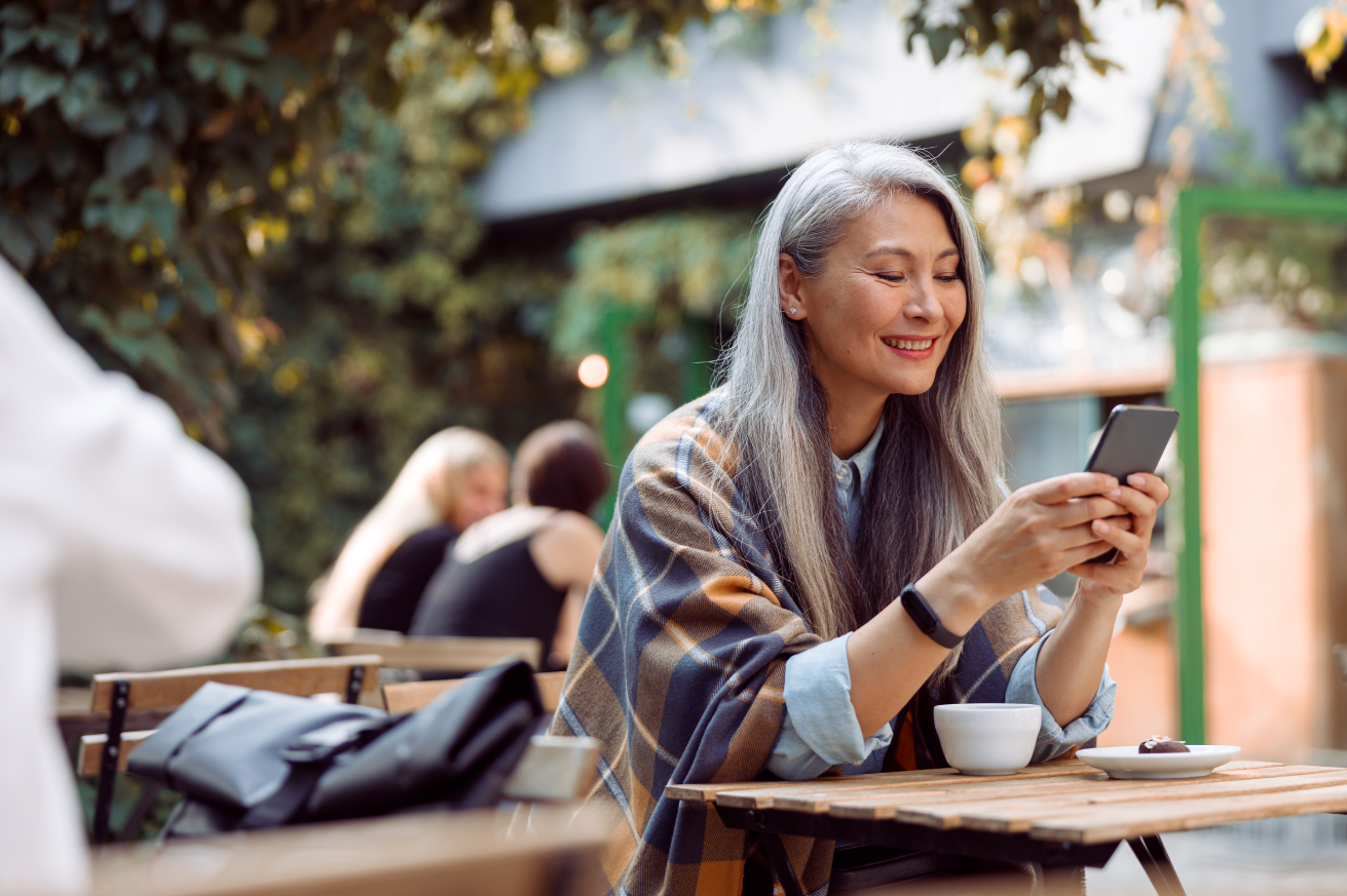 A person with long hair sits at an outdoor café, smiling while looking at a smartphone. A cup is on the table. The backdrop features trees and blurred background figures.
