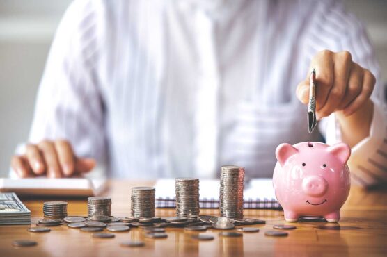 A person putting a dollar bill into a pink piggy bank next to stacks of coins