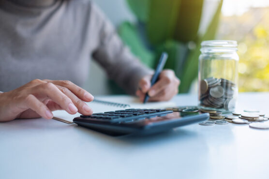Person using a calculator and taking notes next to a jar of coins, representing savings and financial planning