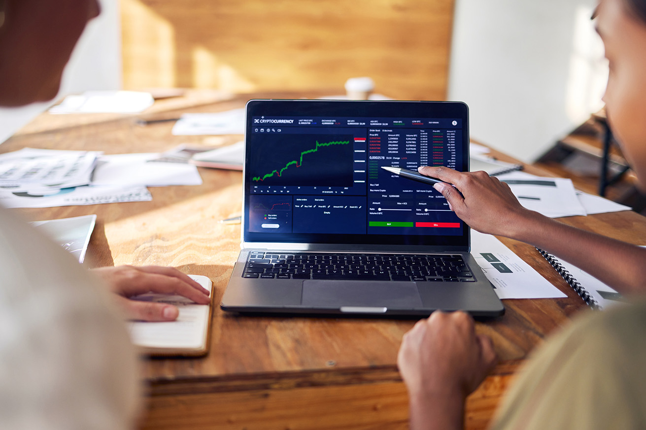 Two people sitting at a wooden table looking at a cryptocurrency trading platform on a laptop