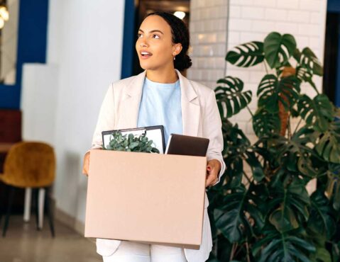 A young woman in a blazer holding a cardboard box filled with office supplies and a plant