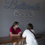 Two young woman sitting at table in front of mural that states "Buttermilk Provisions"