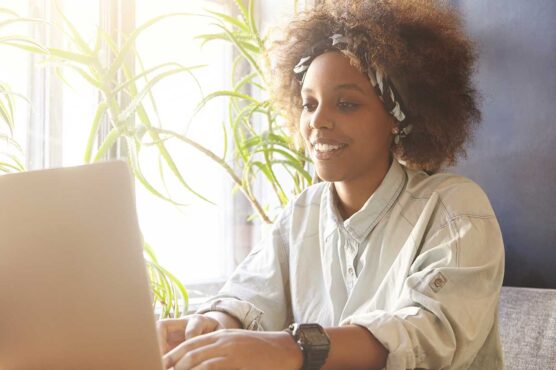 A smiling young woman with an afro and headband working on a laptop near a window