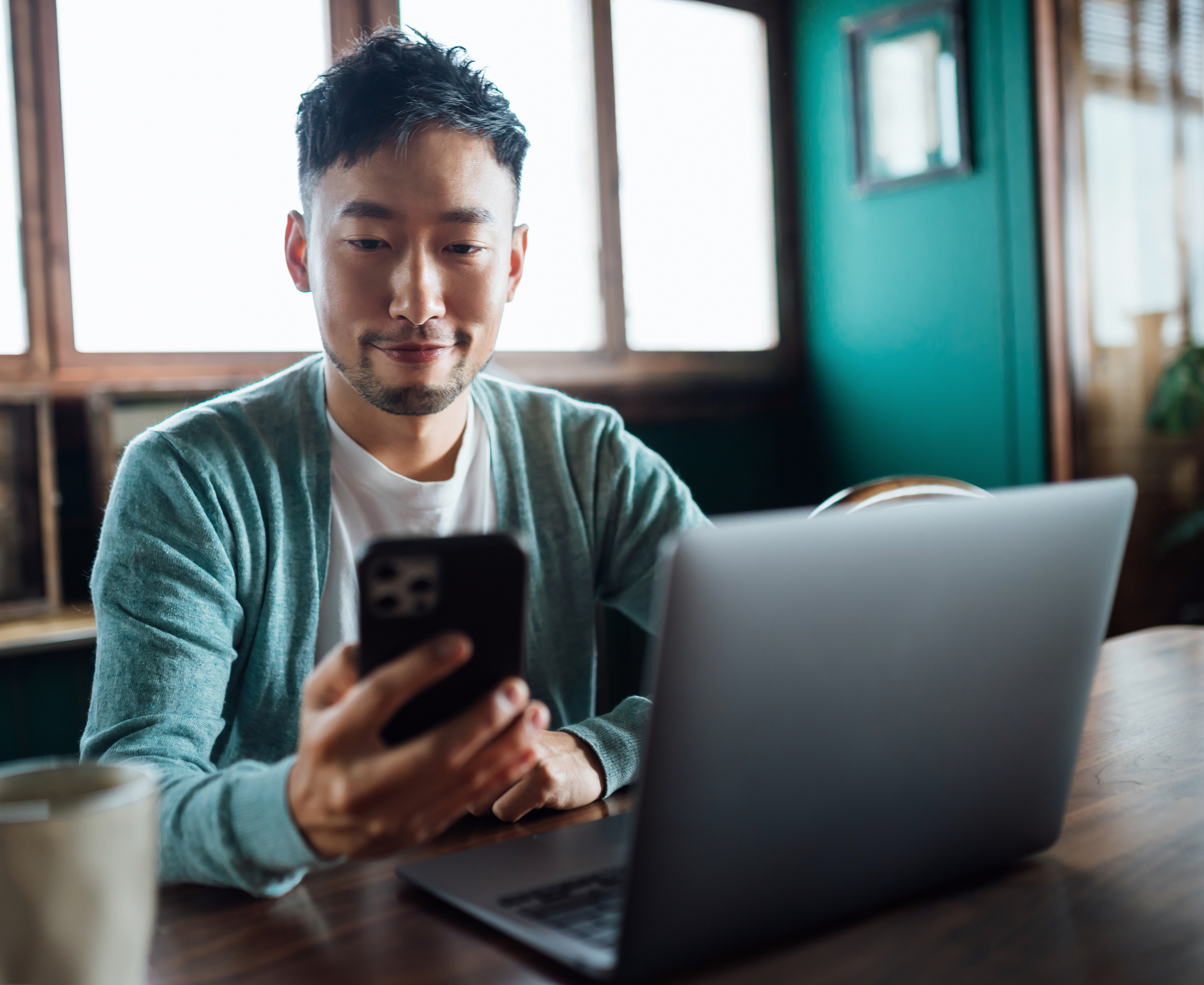 Young man looking at smartphone while working on laptop computer in home office