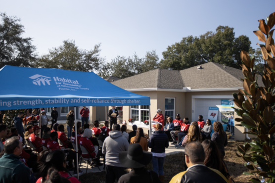 Large group of people under a Habitat for Humanity tent in front of a house listening to a speaker at the Holiday Home Build with the Bucs.