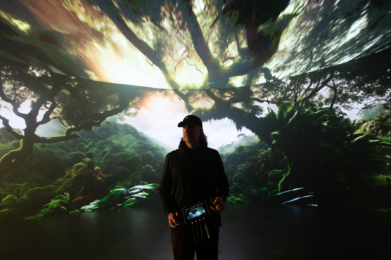 Man holding video production equipment with lush background of green trees and hillsides, water, and sky.