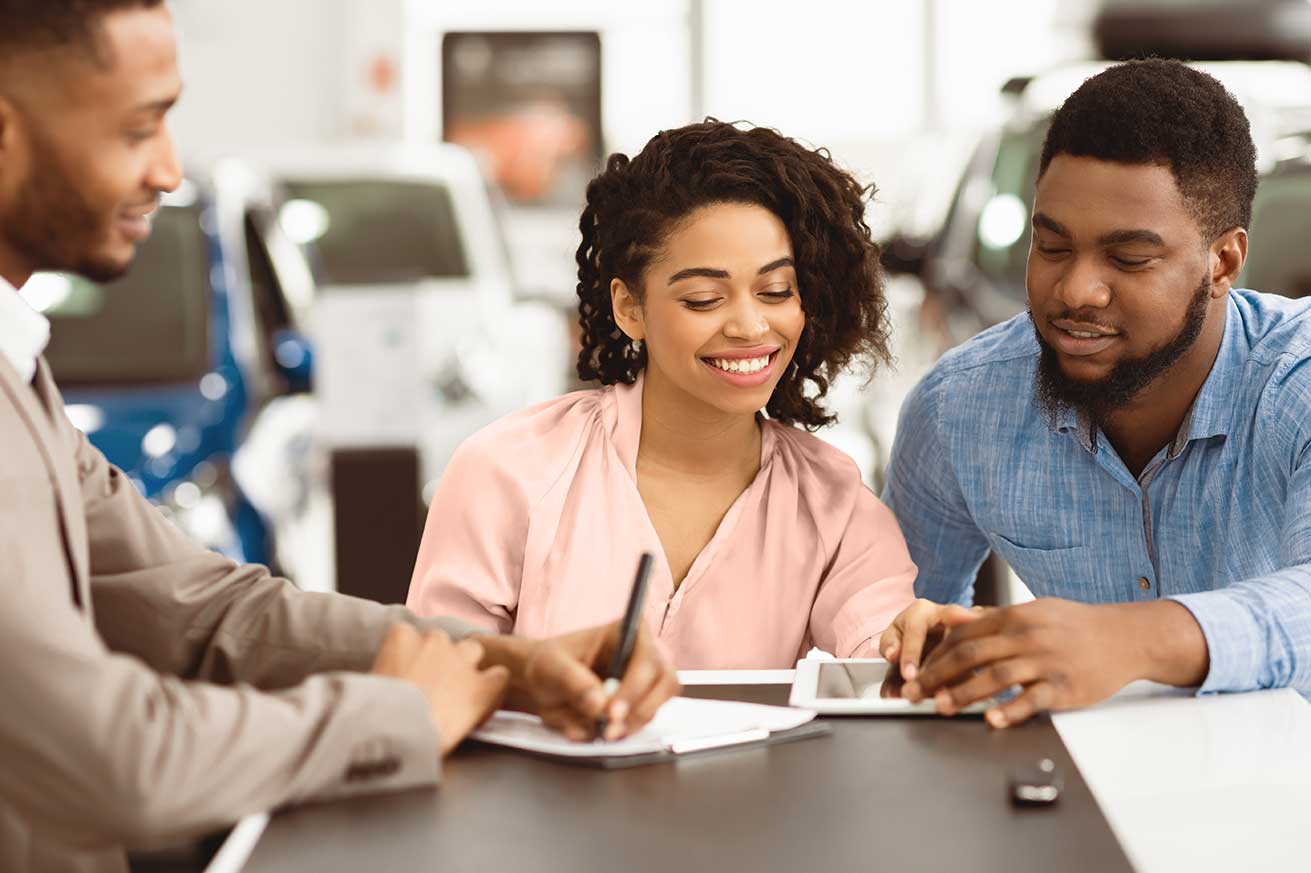 A group of four people are sitting around a table in a car dealership. They appear to be in discussion, with papers and a tablet on the table. Cars are visible in the background through large windows.