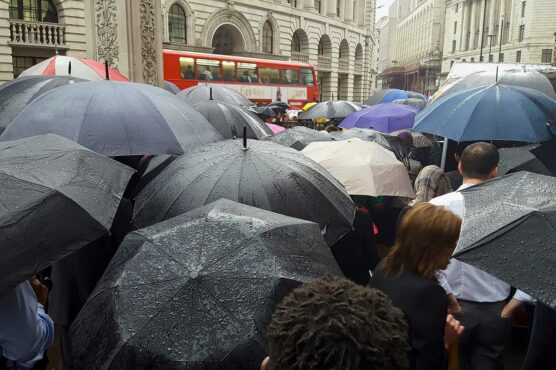 A crowd of people walk with umbrellas on a rainy city street, with a red double‑decker bus in the background.