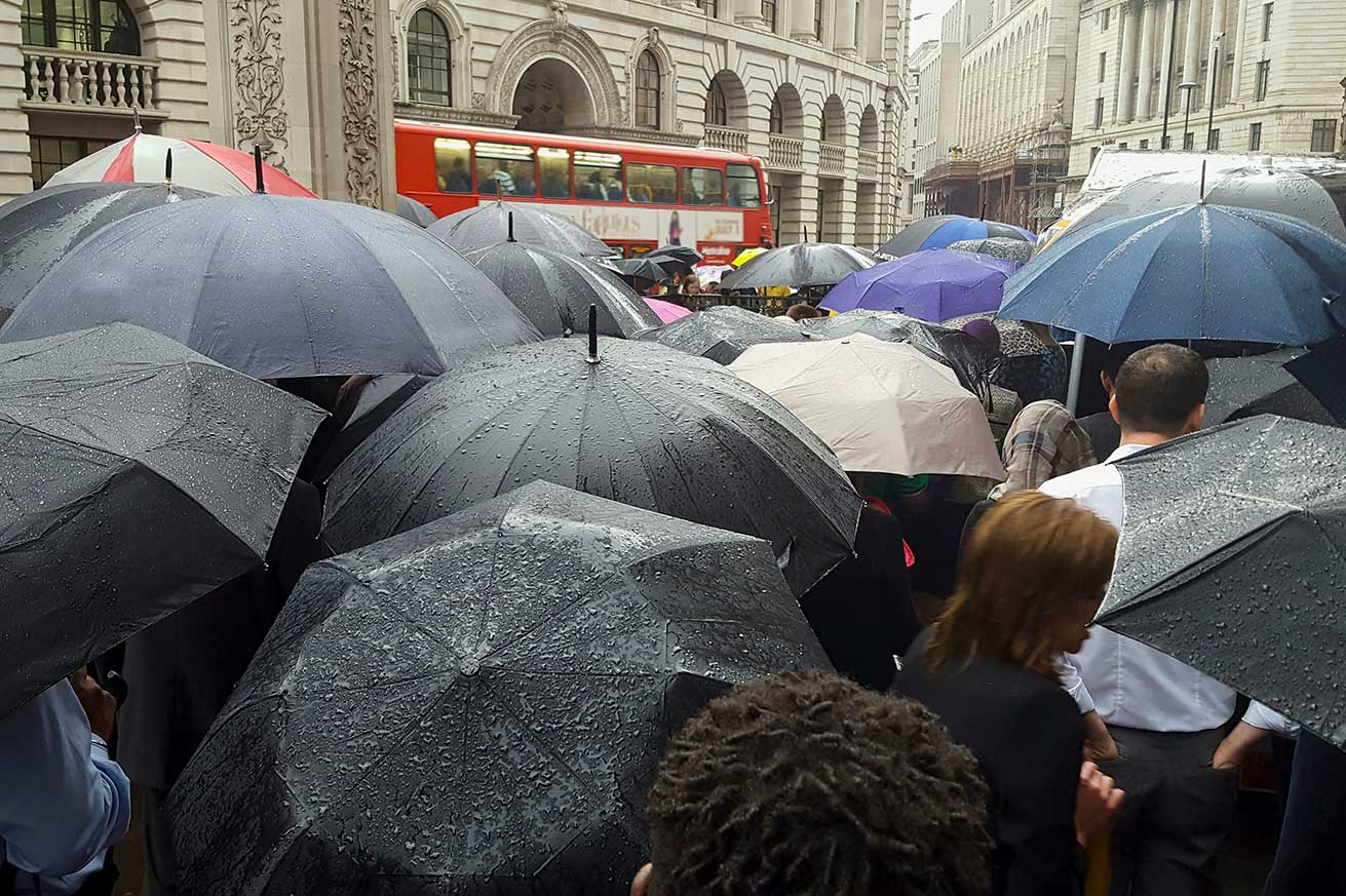 A crowd of people walk with umbrellas on a rainy city street, with a red double‑decker bus in the background.