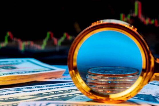A magnifying glass rests over a stack of coins placed on U.S. dollar bills, with a blurred stock market chart and candlestick graph visible in the background, symbolizing investing and financial growth.