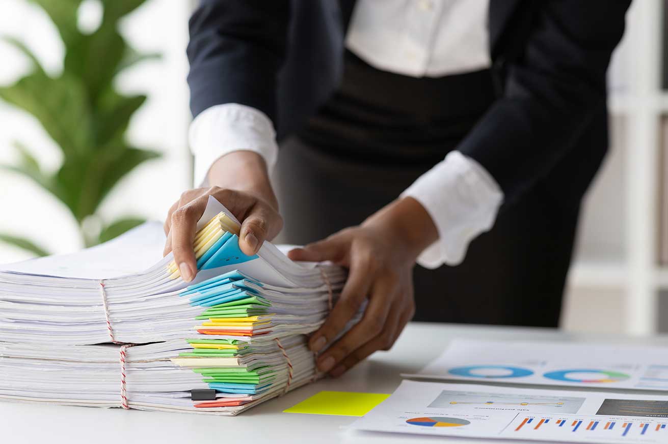Hands organizing a thick stack of documents with colorful tabs on a desk, next to printed charts and graphs.