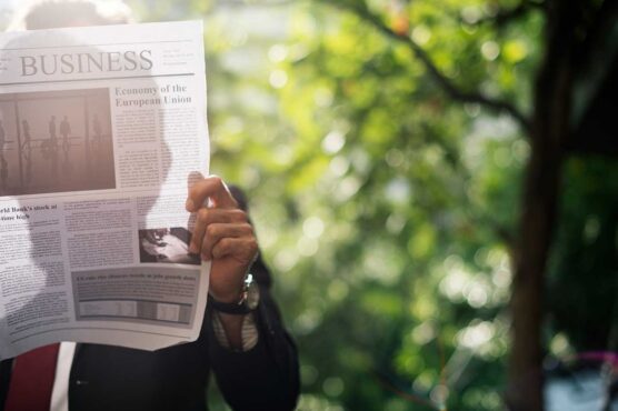 A person holds up a business newspaper open to an article about the European Union economy, with sunlight filtering through green trees in the background.