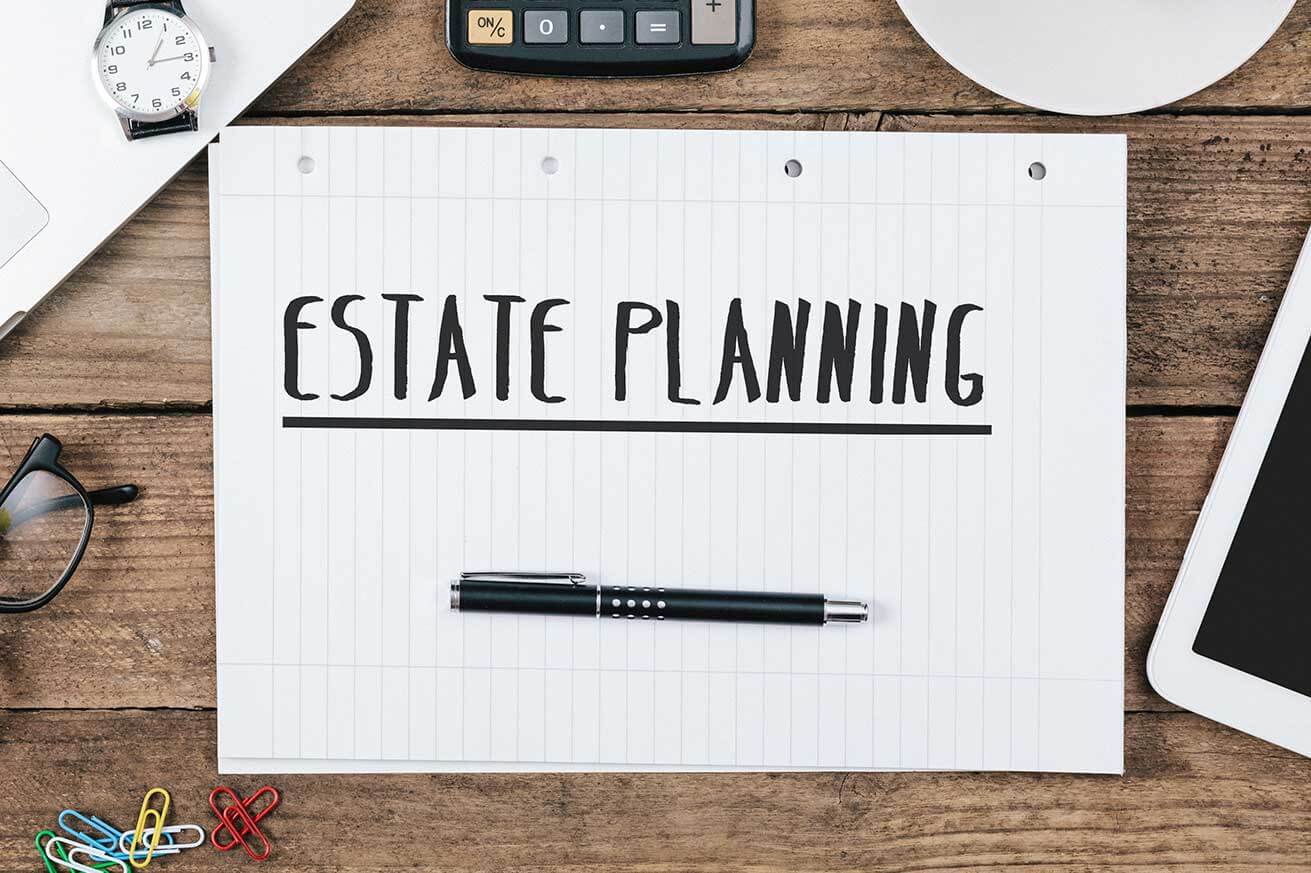 Overhead view of a notebook page labeled ‘Estate Planning’ on a wooden desk, with a pen, calculator, watch, glasses, and paper clips arranged around it.