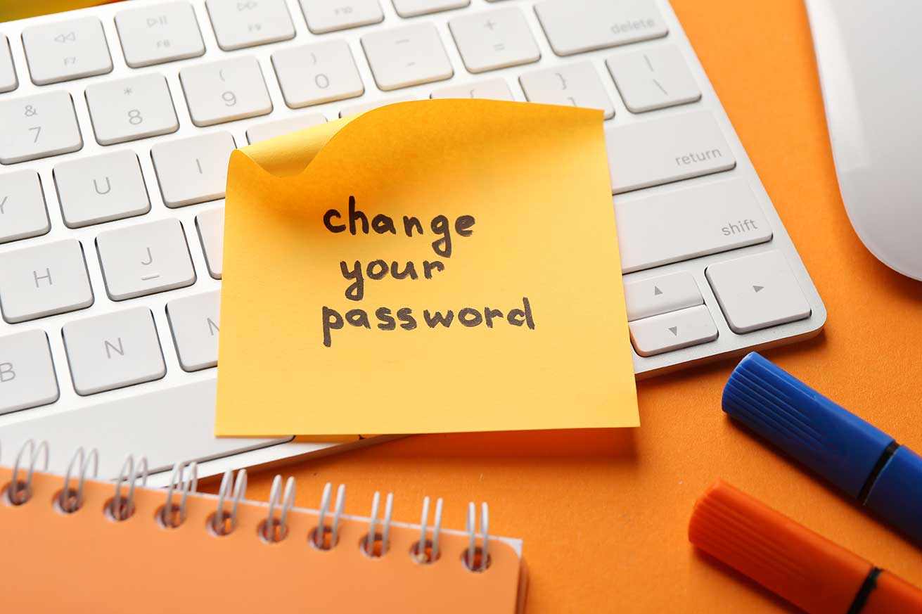 A yellow sticky note reading “change your password” sits on a white computer keyboard on an orange desk, alongside a spiral notebook and colorful markers, emphasizing online security and password safety.