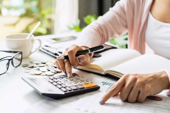 A person sits at a desk using a calculator and reviewing a document with charts and graphs. The workspace includes a notebook, pen, eyeglasses, coins, a cup, and a small potted plant.