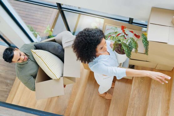 Two people carry boxes and a potted plant into a home, with more moving boxes stacked near a staircase.