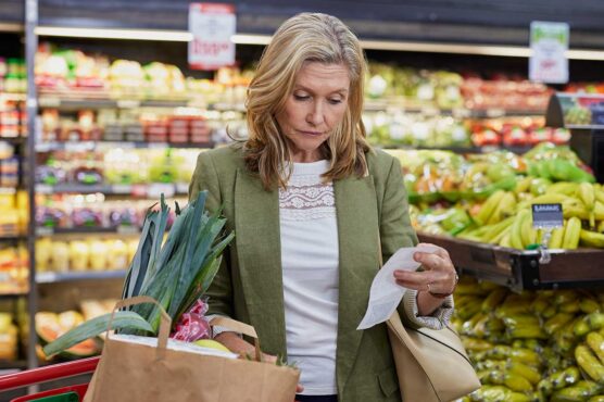 A shopper stands in a grocery store produce aisle holding a paper bag filled with fresh vegetables and fruit while reviewing a printed receipt, with shelves of bananas and other produce in the background.