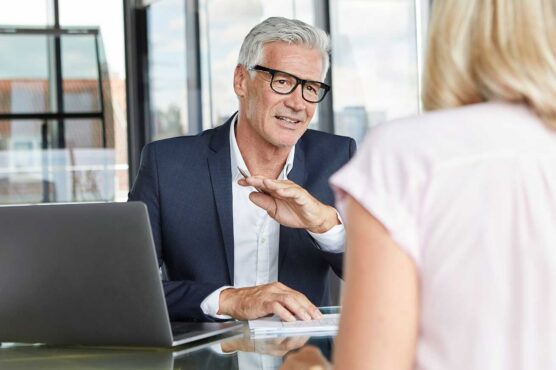 An older man sits at a desk with a laptop, speaking to another person across from him in a bright, modern office.