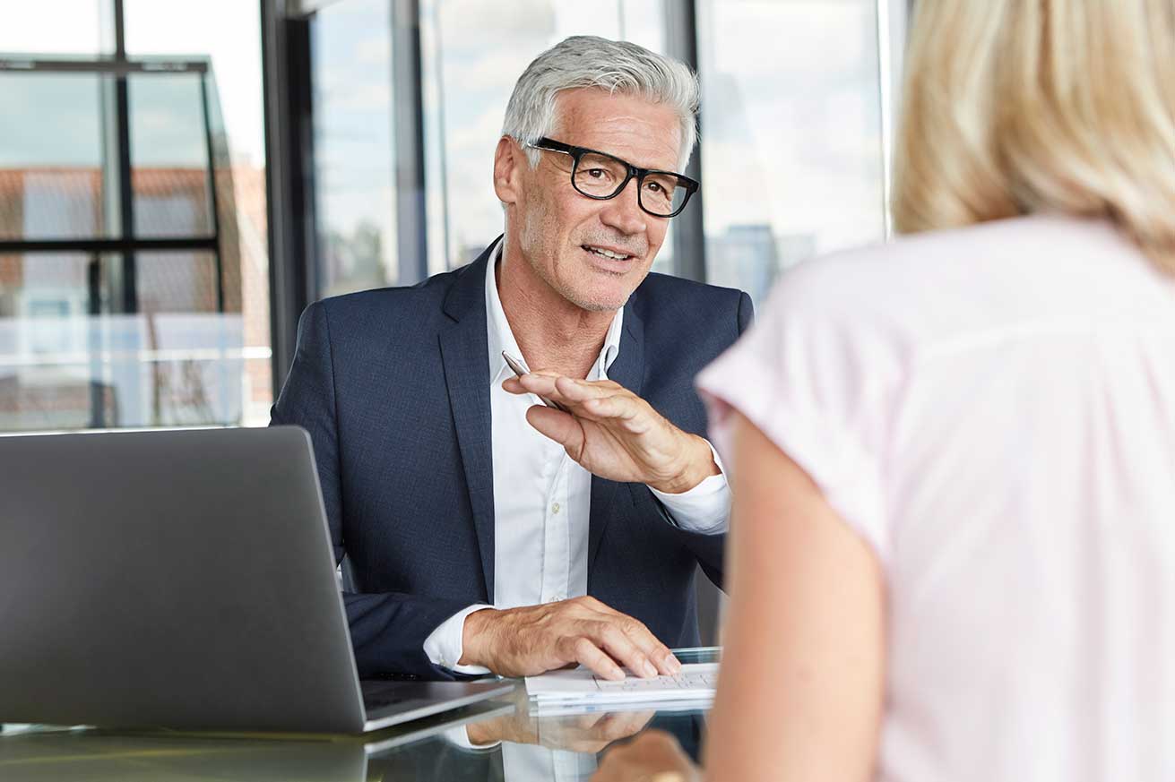 An older man sits at a desk with a laptop, speaking to another person across from him in a bright, modern office.