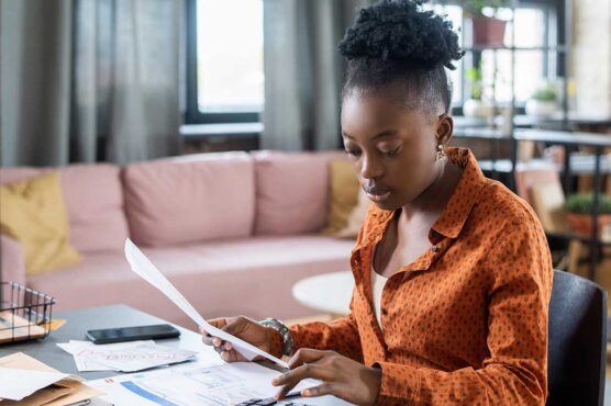 A person sits at a desk in a bright living space, reviewing documents beside a laptop, with a pink couch and indoor plants in the background.