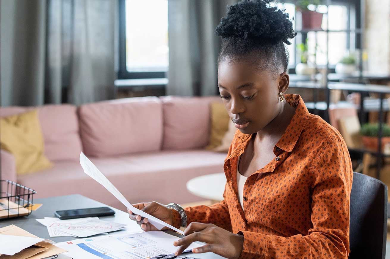 A person sits at a desk in a bright living space, reviewing documents beside a laptop, with a pink couch and indoor plants in the background.