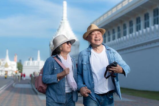 Two adults wearing casual denim outfits and sun hats walk along a paved path beside an ornate white building, with one holding a camera, under a clear blue sky.