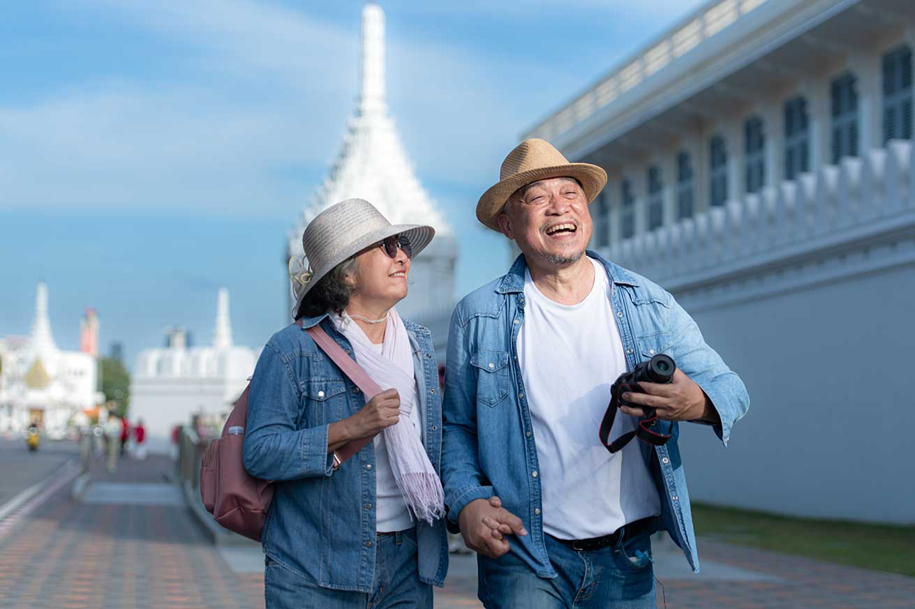 Two adults wearing casual denim outfits and sun hats walk along a paved path beside an ornate white building, with one holding a camera, under a clear blue sky.