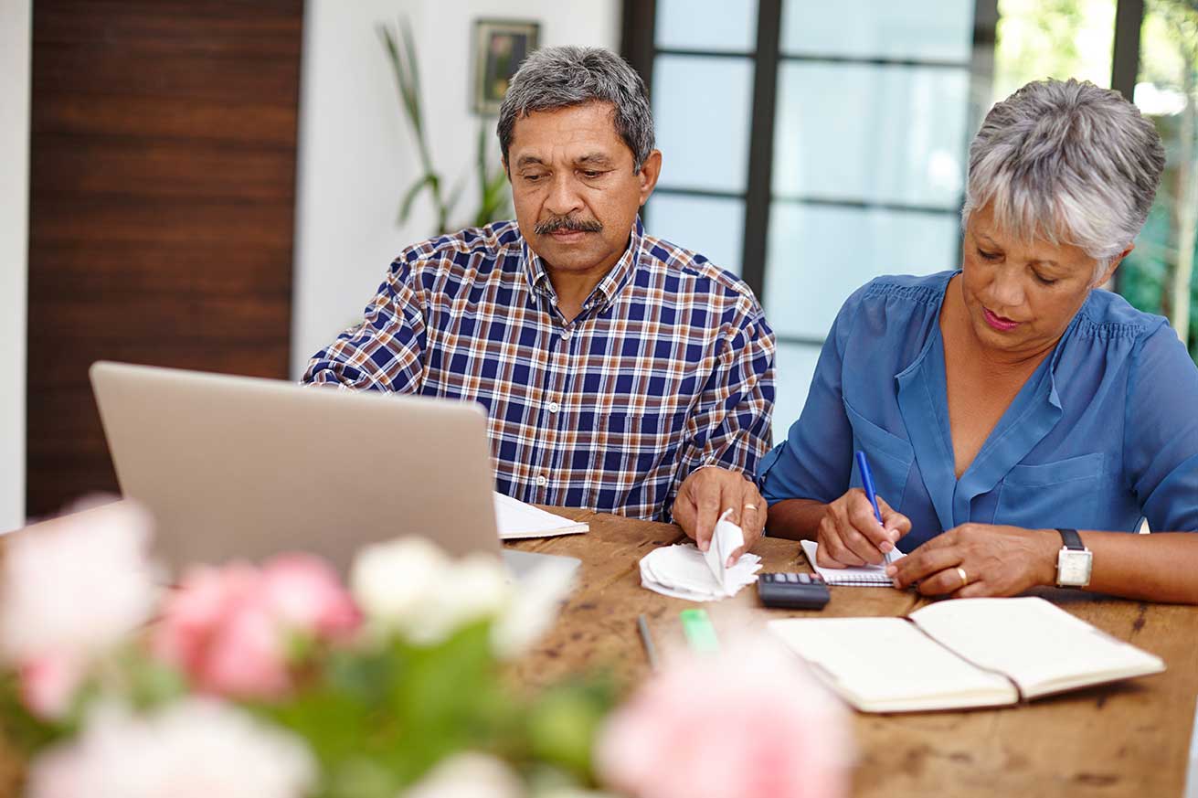 Two adults sitting at a table reviewing financial documents, using a laptop and calculator while taking notes.