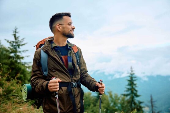 A person stands outdoors with a backpack and trekking poles, looking out over misty green mountains and trees.
