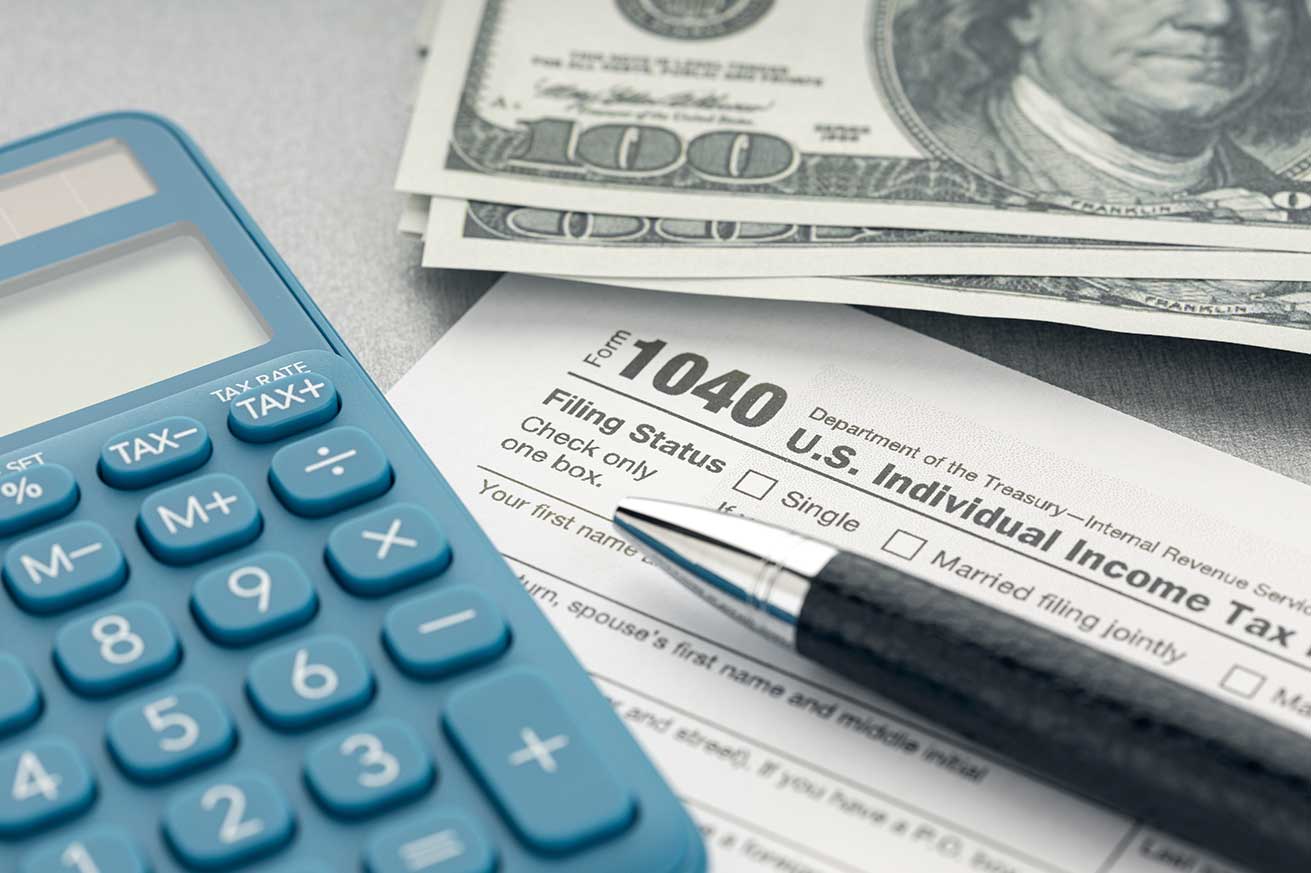 A calculator, pen, stack of hundred‑dollar bills, and a partially completed IRS Form 1040 are arranged on a desk.