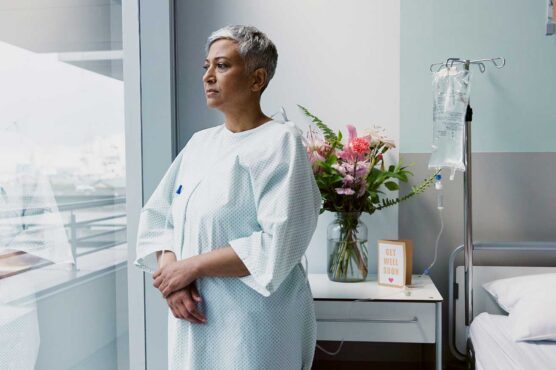 A patient in a light blue hospital gown stands by a large window in a hospital room, with an IV stand nearby and a bedside table holding a vase of flowers and a “Get Well Soon” card.