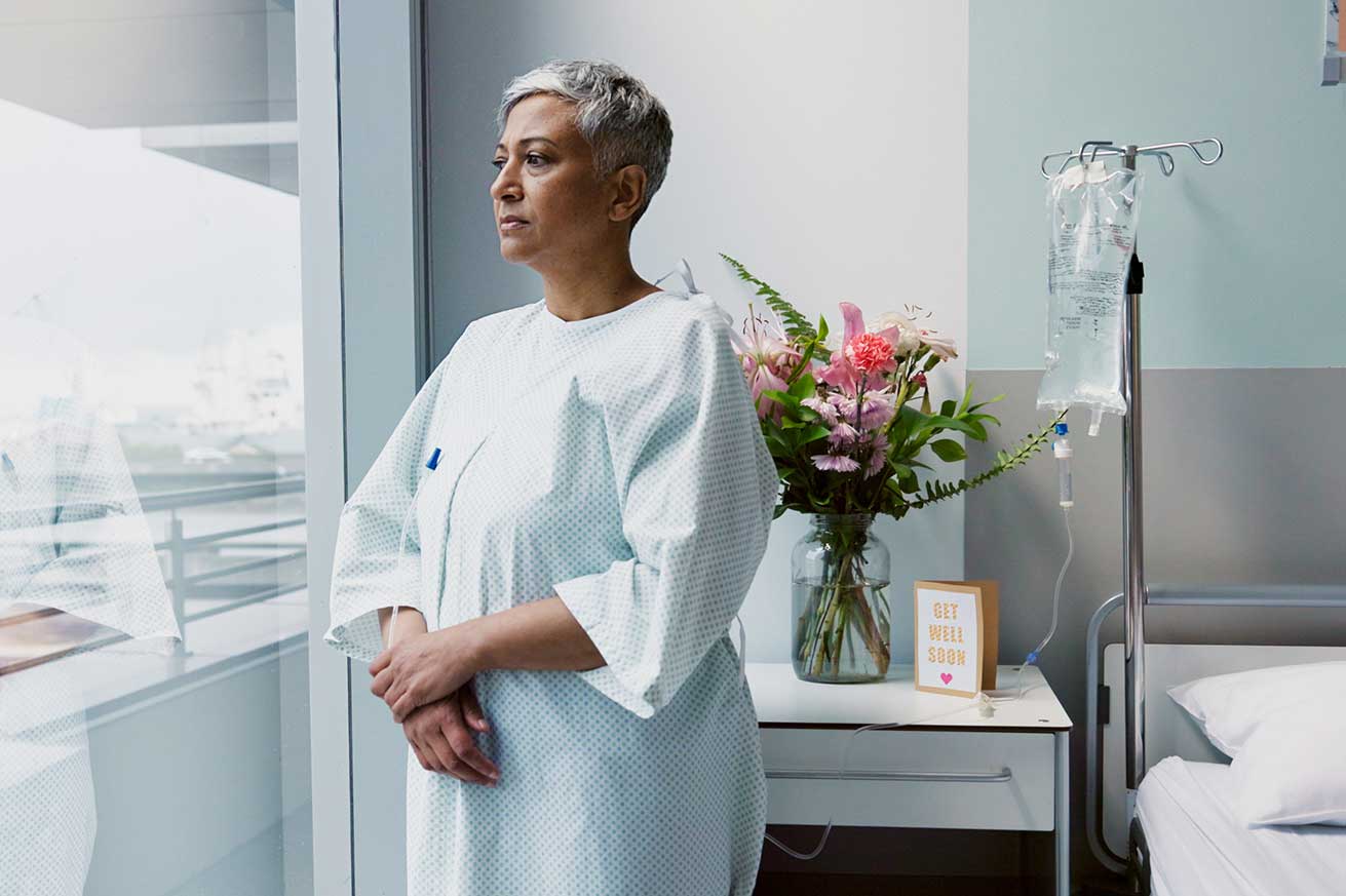A patient in a light blue hospital gown stands by a large window in a hospital room, with an IV stand nearby and a bedside table holding a vase of flowers and a “Get Well Soon” card.