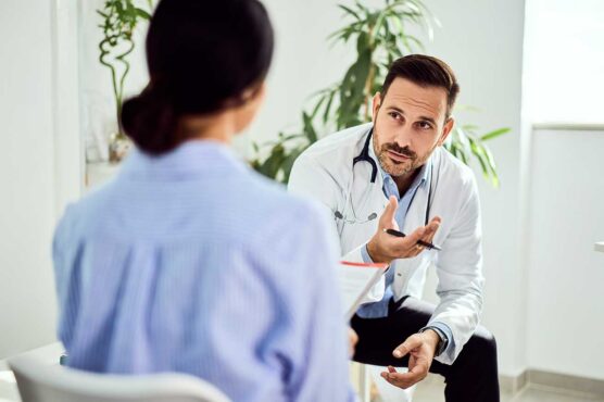 A doctor in a white coat with a stethoscope speaks with a patient across a desk, holding papers in a bright medical office with plants in the background.