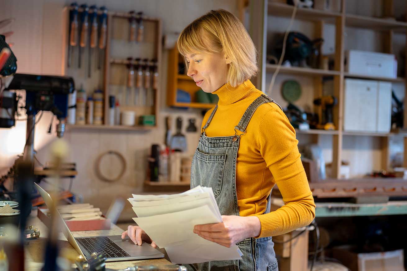 A person in a woodworking studio looks at a laptop while holding papers, surrounded by tools, shelves, and equipment.