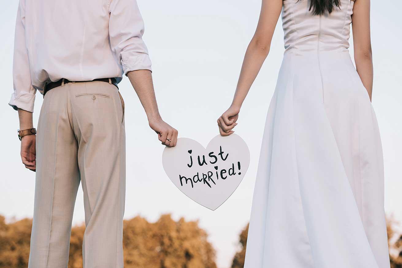 A couple stands outdoors holding a white heart‑shaped sign that reads “just married,” shown from behind with trees and sky in the background.