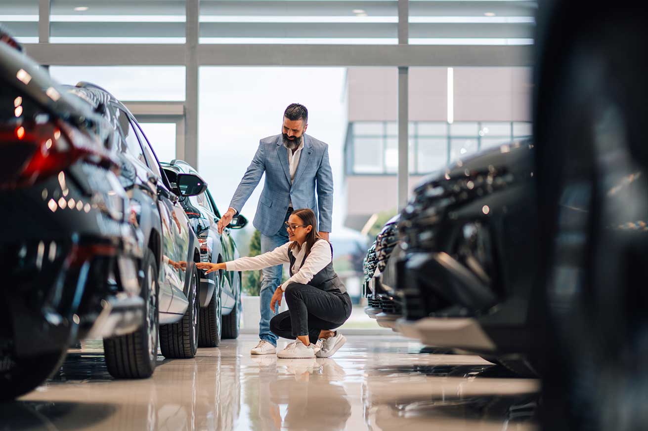 Two people stand inside a bright car showroom. One person crouches beside a parked car, examining the side, while the other stands nearby pointing toward the vehicle. Several cars line the polished floor, and large windows let in natural light.