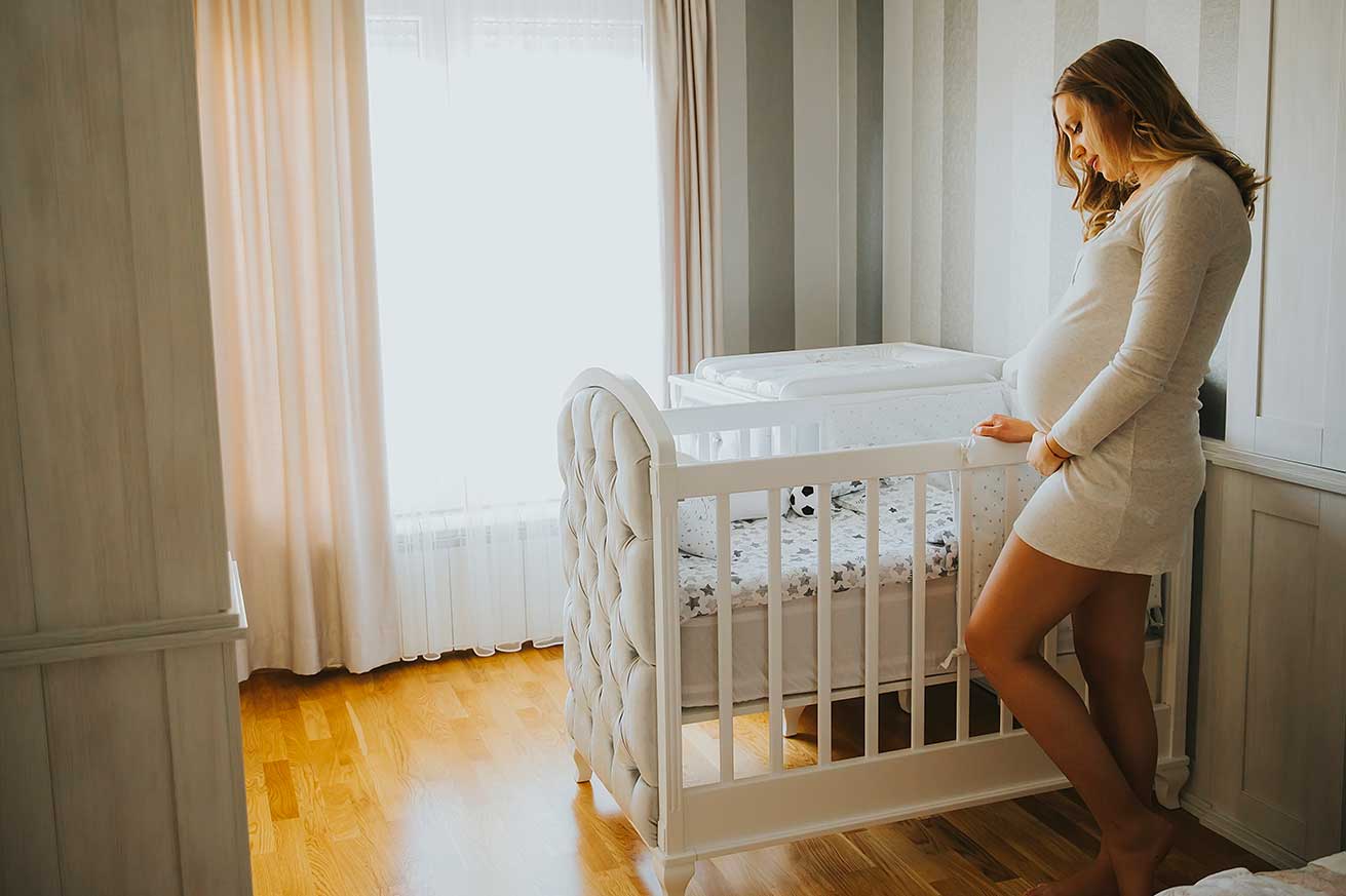 A pregnant person stands beside a white crib in a softly lit nursery with striped walls, sheer curtains, and wooden flooring. The crib has tufted ends and patterned bedding with a small stuffed toy.