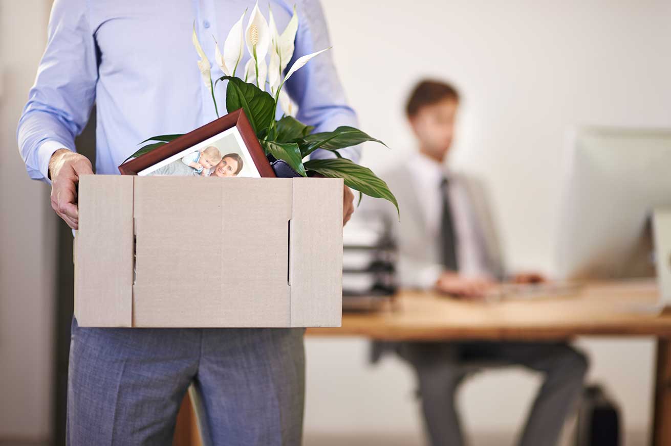 A person holds a cardboard box filled with personal items, including a framed photo and a potted plant, while another person works at a computer in the background of an office.