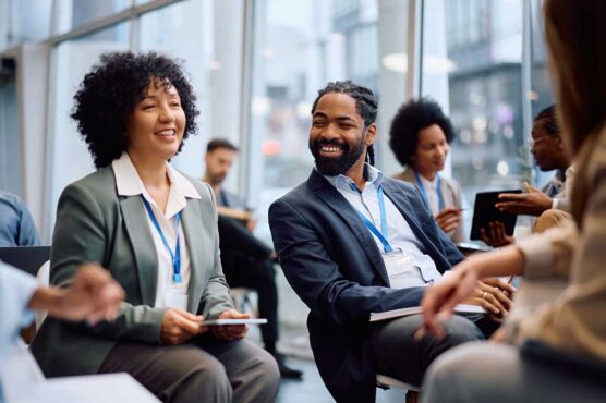 A group of people wearing business attire sit together with notebooks and badges, talking in a bright conference space with large windows and city buildings in the background.