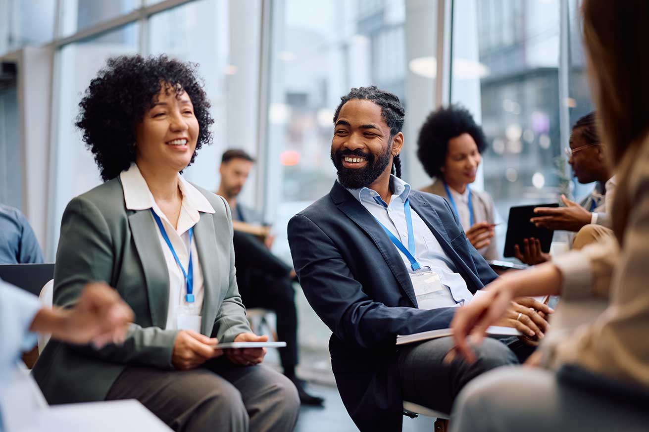 A group of people wearing business attire sit together with notebooks and badges, talking in a bright conference space with large windows and city buildings in the background.