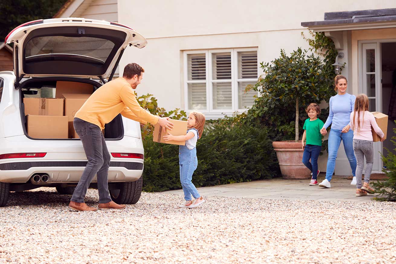 A family stands outside a house loading or unloading cardboard boxes. A white SUV with its back hatch open sits in the driveway, filled with boxes. An adult hands a box to a child near the car, while another adult and two children walk from the house carrying more boxes.