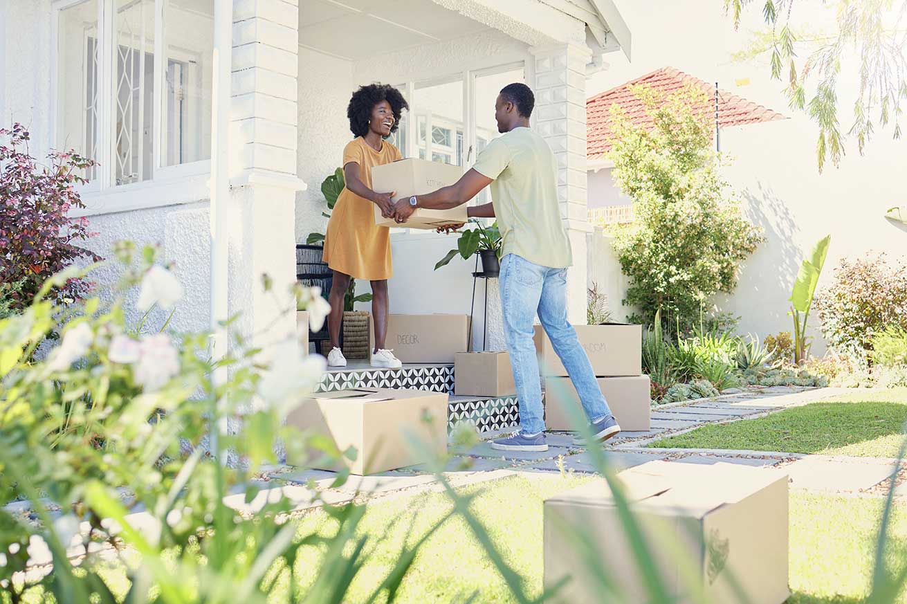 Two people stand outside a white house surrounded by greenery, passing a cardboard box between them on the front walkway. Several labeled moving boxes sit nearby, suggesting they are moving in or out.