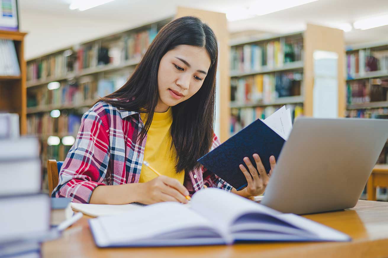 A person sits at a desk in a library, reading a book while writing in a notebook. A laptop is open in front of them, and several stacks of books sit on the desk. Bookshelves filled with books line the background.