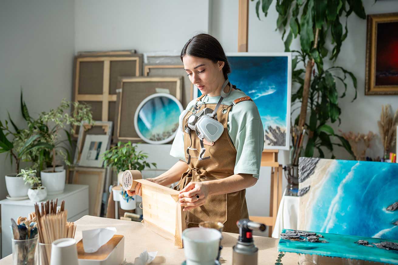 An artist in a studio works on a wooden panel while wearing an apron, surrounded by brushes, cups, and ocean‑themed paintings in the background.