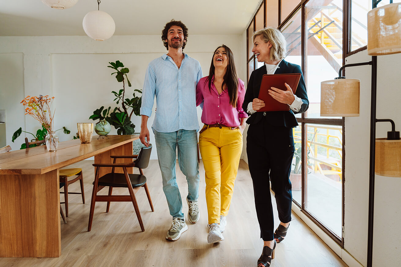 Three people walk through a bright, modern home with large windows and wooden floors. One person holds a folder, appearing to guide the other two as they tour the space. A long dining table with chairs, plants, and decor sits to the side.