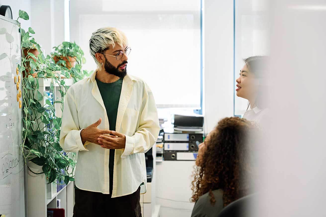 A person stands at a whiteboard explaining a concept while two colleagues listen, in a modern office with plants and shelves in the background.
