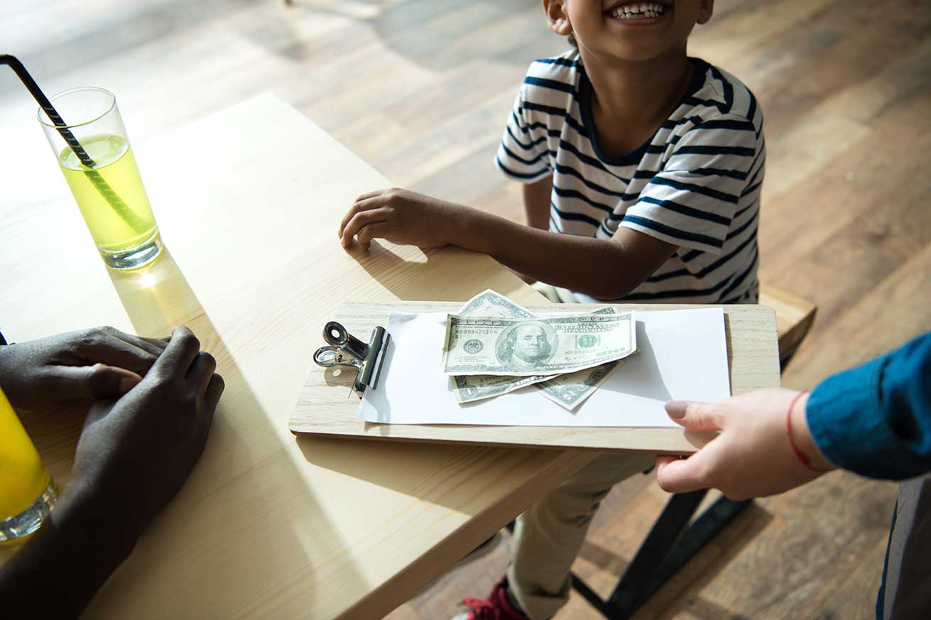 A person hands a clipboard with a receipt and cash to two people seated at a wooden table, including a child in a striped shirt and another person with a yellow-green drink.