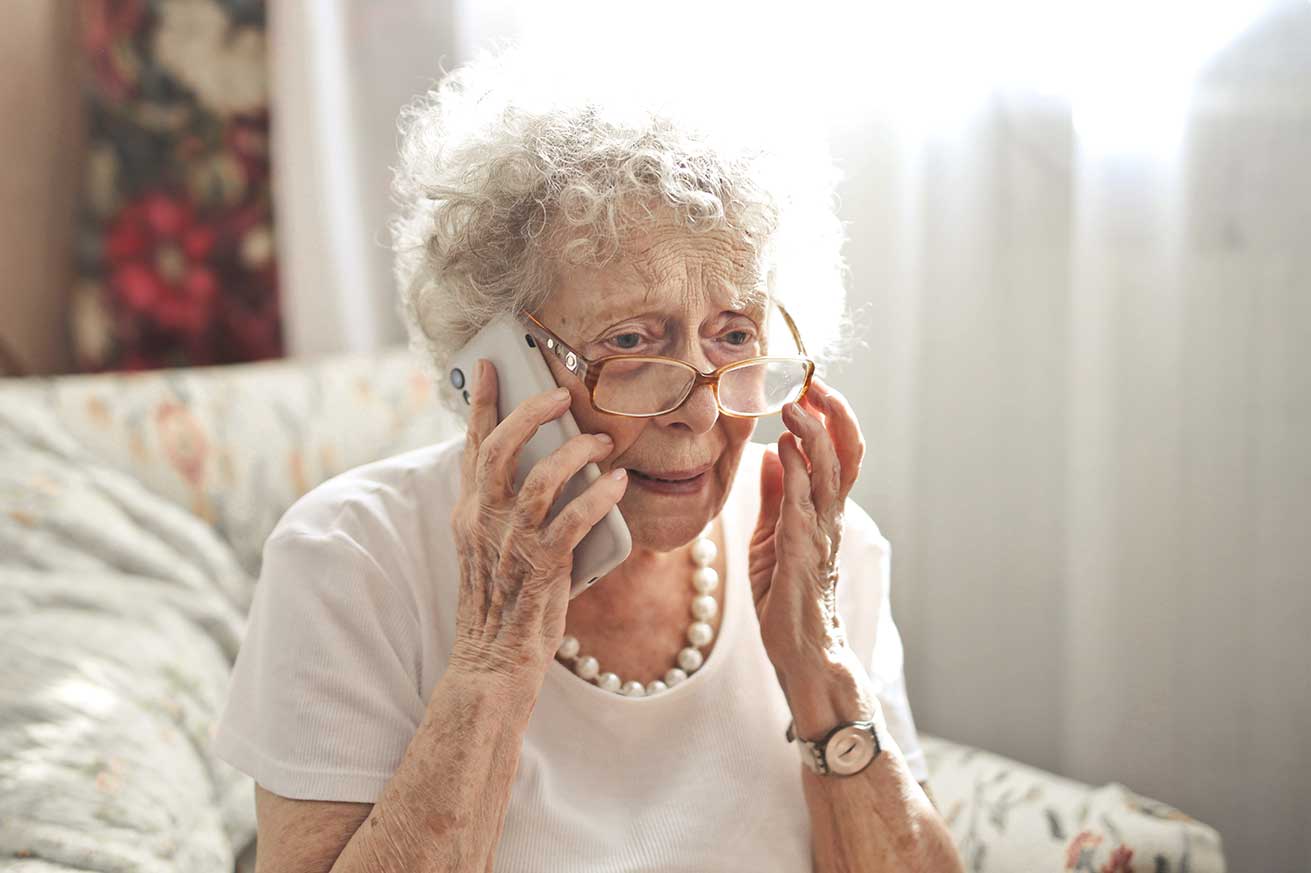An older person with curly gray hair and glasses sits on a patterned couch, holding a smartphone to their ear with one hand while touching their face with the other, softly lit by natural light from nearby curtains.