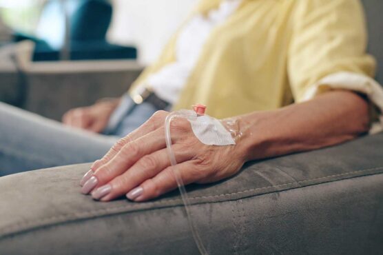 A close-up of a person’s hand resting on a gray armrest with an IV catheter taped to the back of the hand, connected to a clear tube. The person is wearing a yellow shirt and blue jeans, and the background is softly blurred.