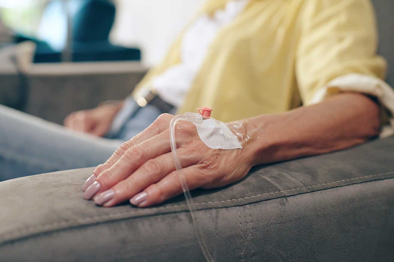 A close-up of a person’s hand resting on a gray armrest with an IV catheter taped to the back of the hand, connected to a clear tube. The person is wearing a yellow shirt and blue jeans, and the background is softly blurred.